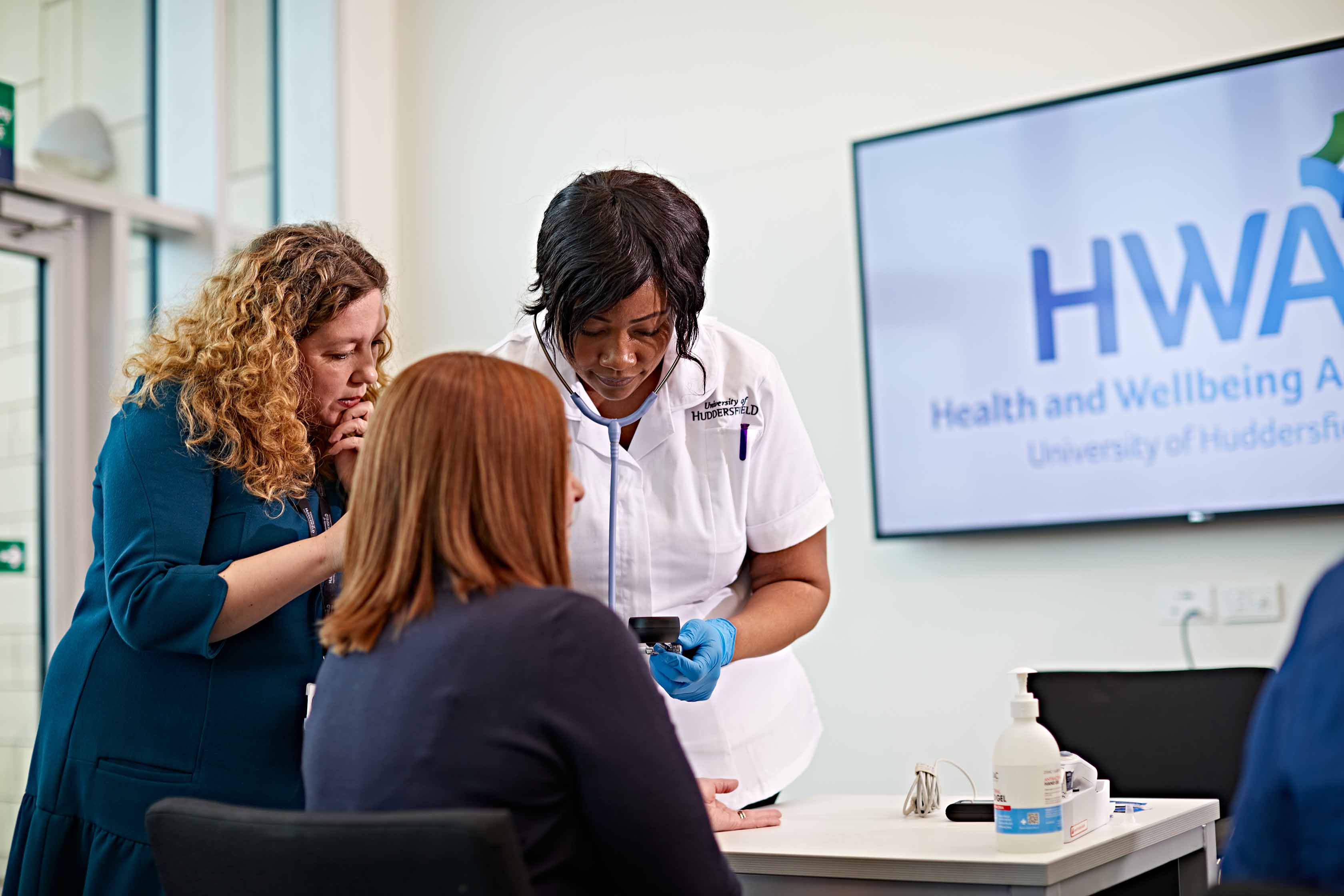 A seated woman is receiving medical attention from a woman in a University of Huddersfield nurses uniform